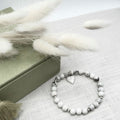 A beaded stretch bracelet with white and grey marbled howlite beads, a silver heart charm, and small silver spacer beads, displayed on a textured light fabric next to a green box and white pampas grass.
