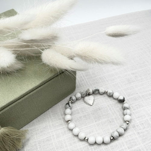 A beaded stretch bracelet with white and grey marbled howlite beads, a silver heart charm, and small silver spacer beads, displayed on a textured light fabric next to a green box and white pampas grass.