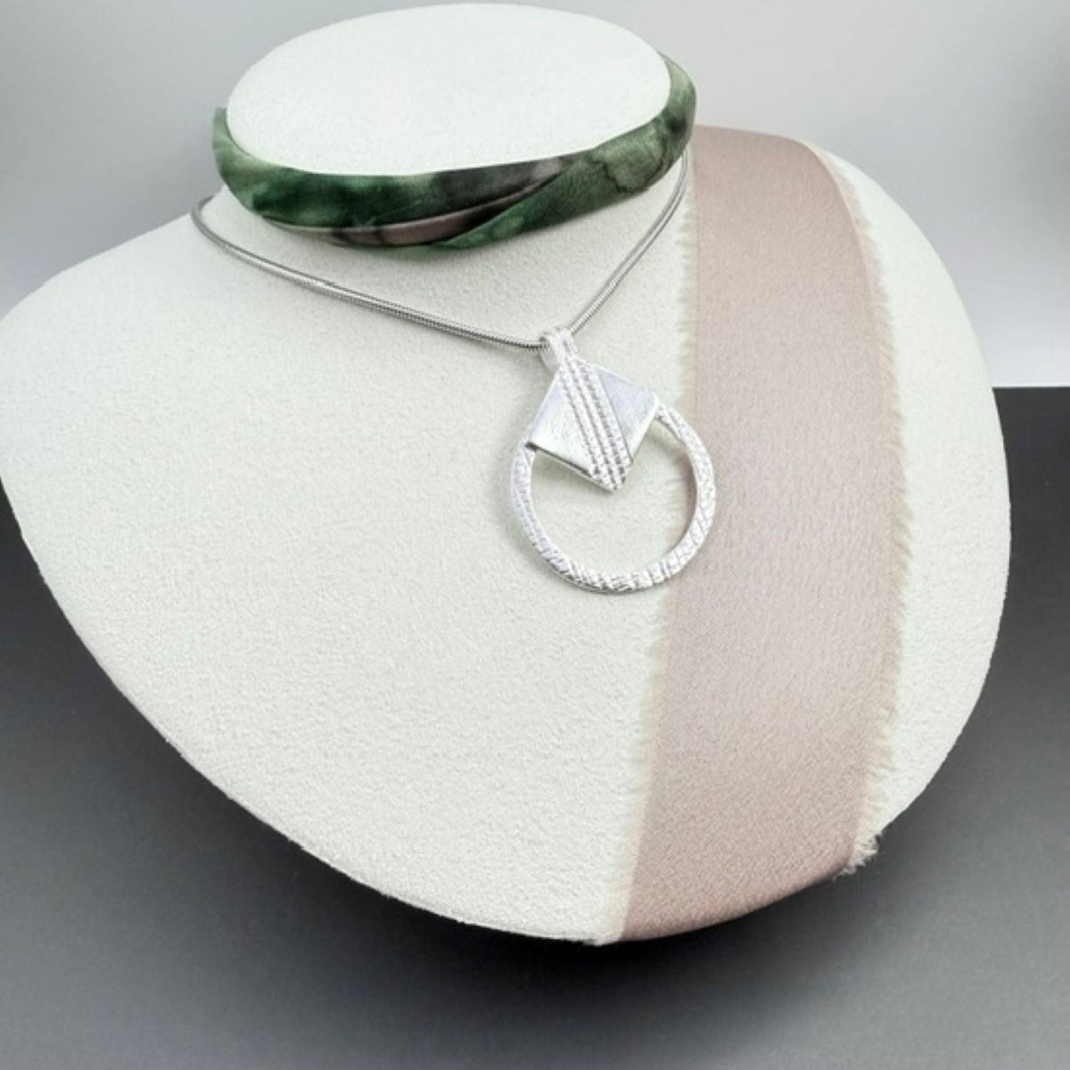 A silver pendant necklace displayed on a white jewellery bust. The necklace shows a textured silver pendant with a geometric square design feature on a textured circular ring paired with a thin silver chain.