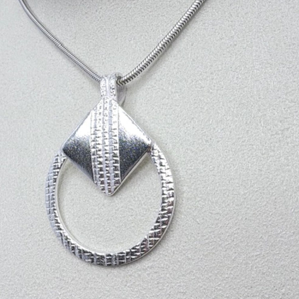 A circular textured silver pendant necklace featuring a silver tilted square on a silver chain, displayed on a textured light grey surface.