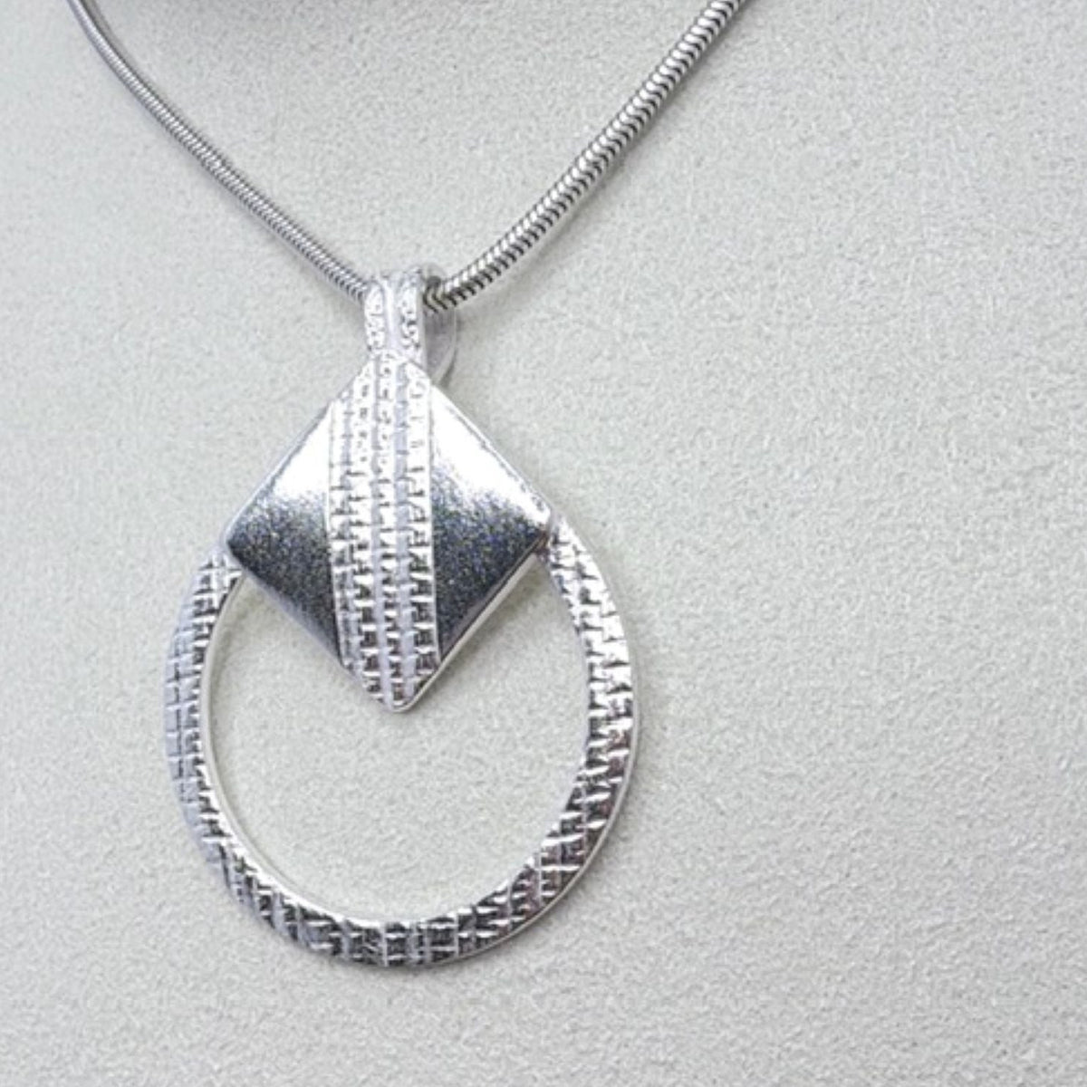 A circular textured silver pendant necklace featuring a silver tilted square on a silver chain, displayed on a textured light grey surface.
