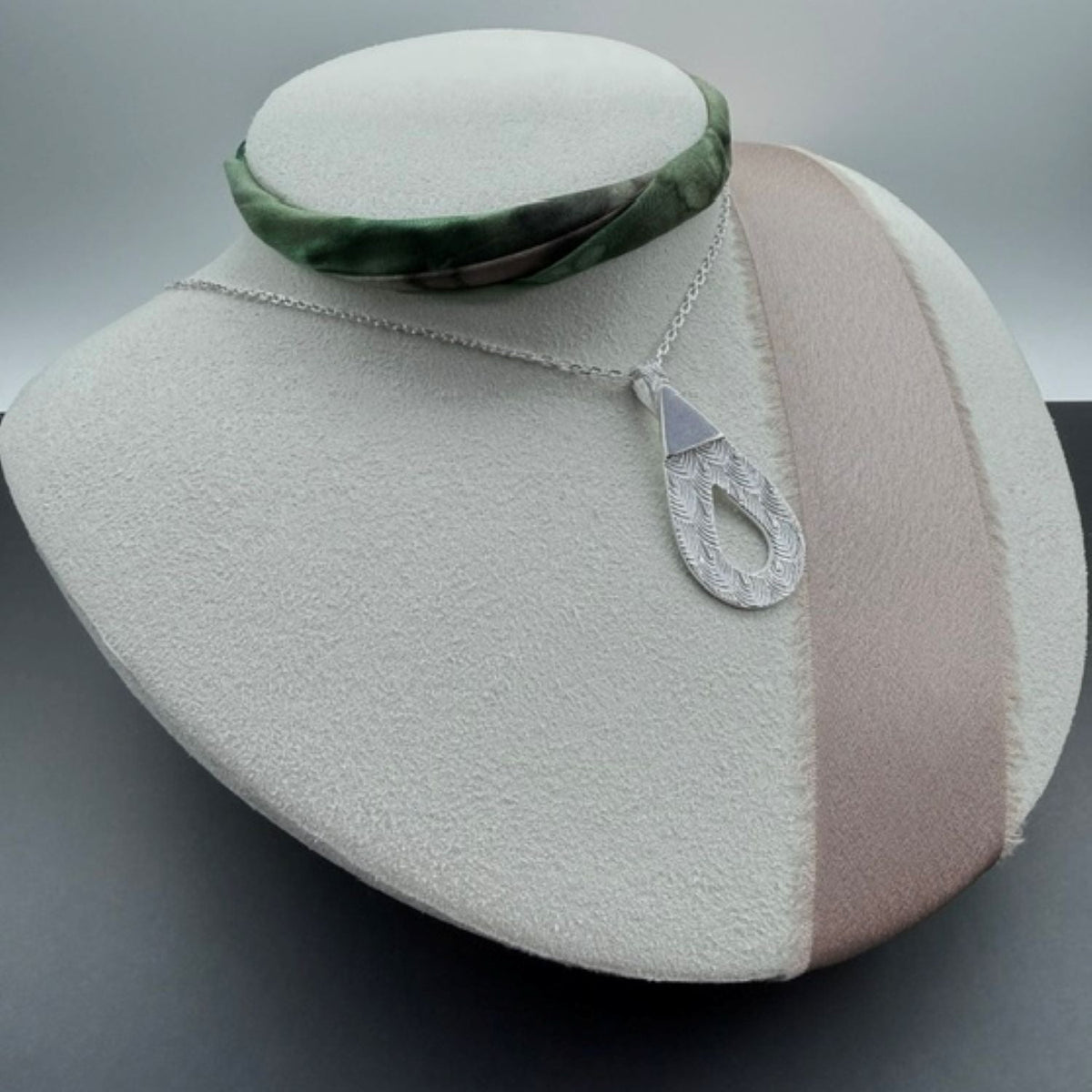 A silver pendant necklace with a textured teardrop-shaped pendant and a matching chain, displayed on a cream-colored jewelry bust. A green and black patterned collar is draped behind the bust.