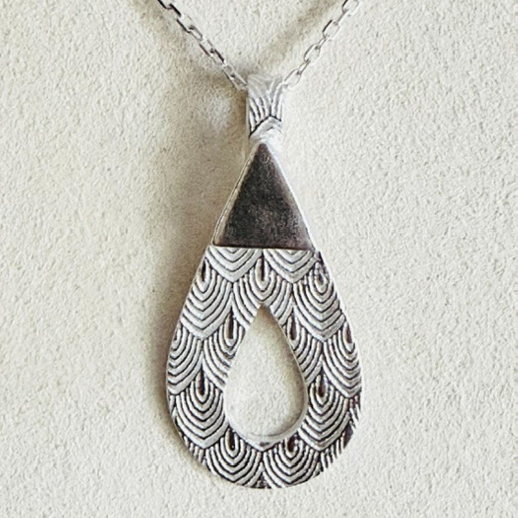 A silver teardrop pendant necklace with a textured scallop pattern and a triangular accent at the top, hanging from a silver chain against a textured off-white background.