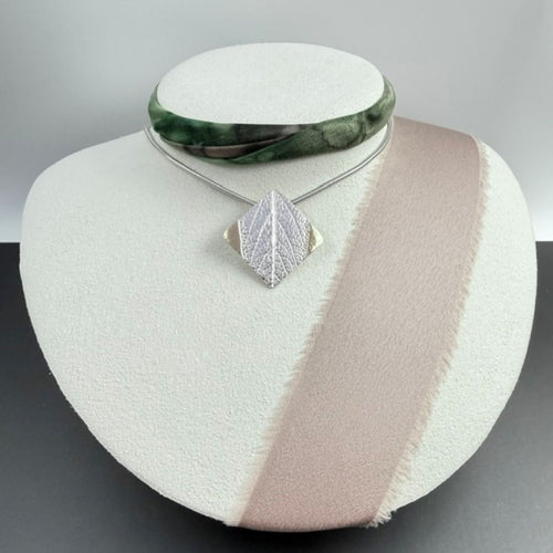 A silver tilted square pendant necklace with a delicate leaf texture and added gold, displayed on a white jewellery bust with a pink ribbon, alongside a green bangle bracelet