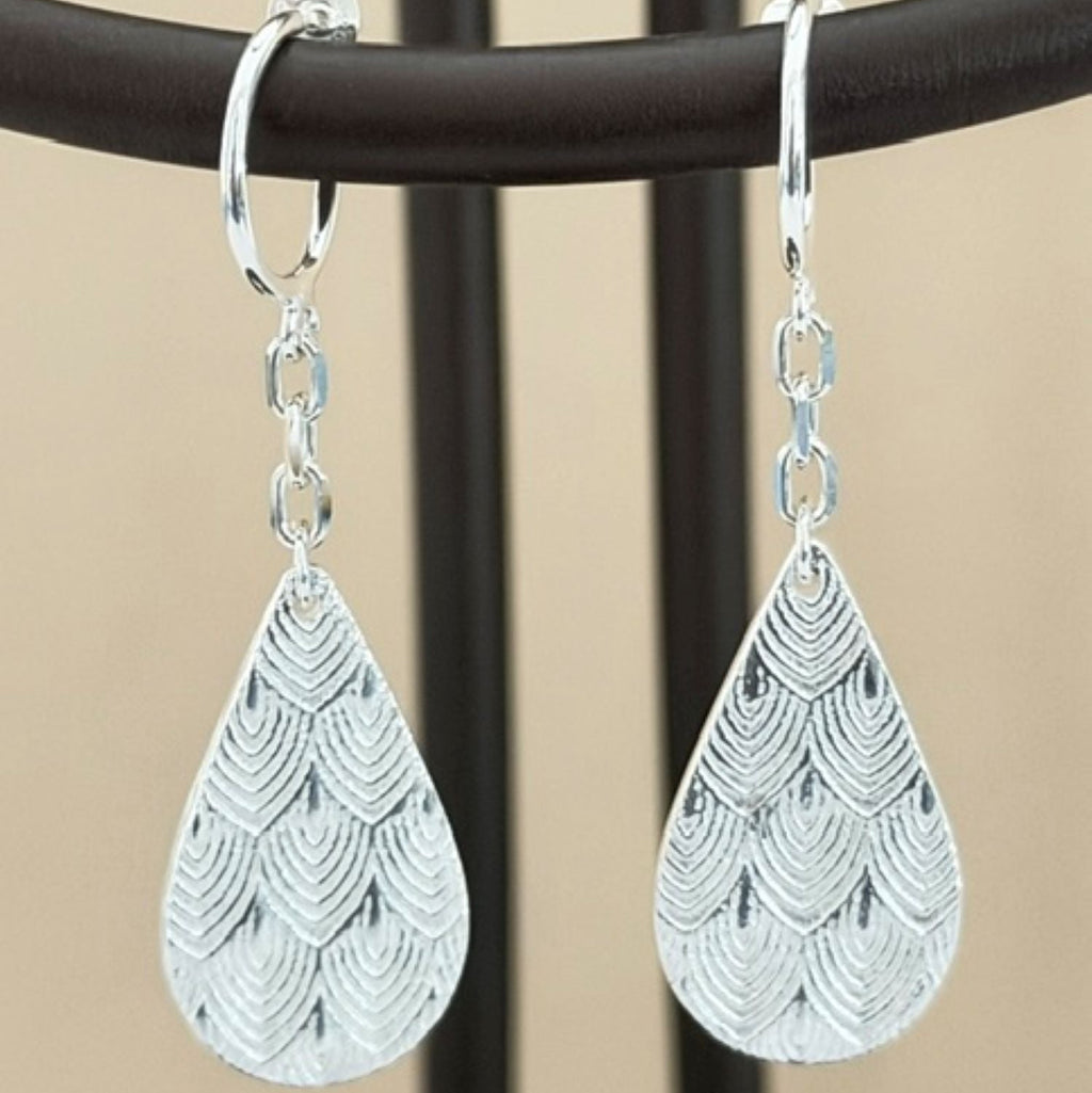 A pair of silver teardrop dangle earrings with a textured scallop pattern, hanging from a black display stand against a beige background