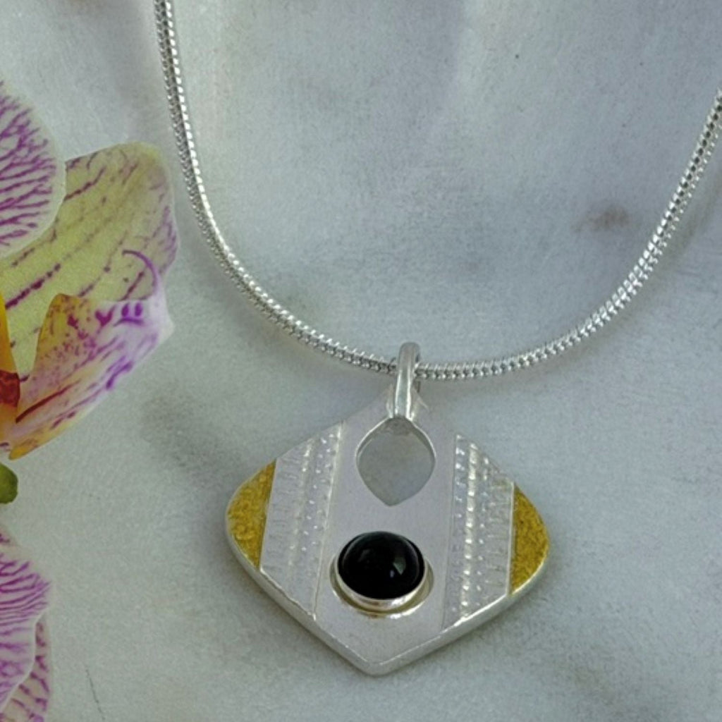 A silver drop-shaped pendant necklace featuring a central black onyx gemstone, textured silver stripes, and gold accents, on a silver chain displayed on a light marble surface with a purple orchid flower on the left.