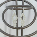 A pair of silver dangle earrings with a textured, geometric diamond design hanging from a dark brown metal display stand.