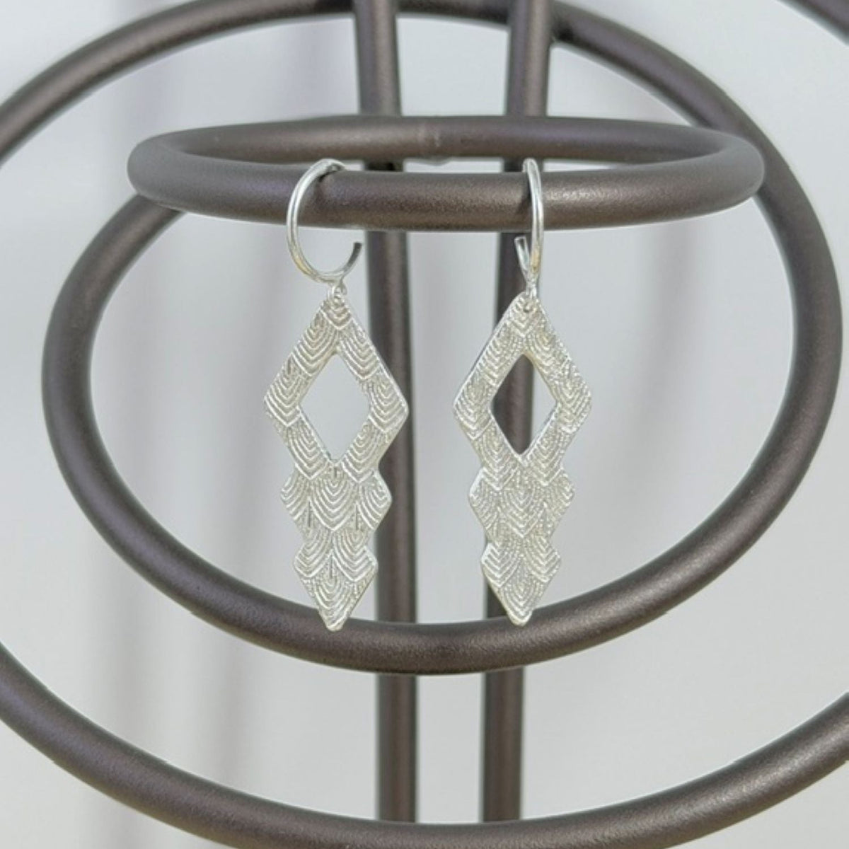 A pair of silver dangle earrings with a textured, geometric diamond design hanging from a dark brown metal display stand.