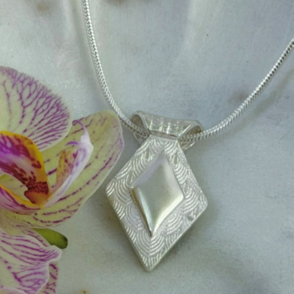 A silver textured diamond-shaped pendant necklace with a central raised diamond shaped plain silver feature, displayed on a light marble surface next to a purple and yellow orchid flower.