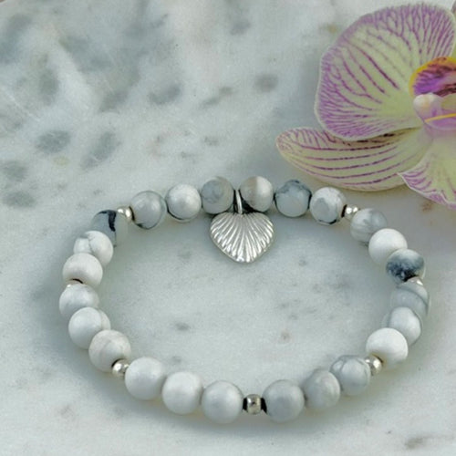 A beaded stretch bracelet featuring marbled white and light grey howlite beads, small silver spacer beads, and a silver heart charm, displayed on a white marble tray next to a purple orchid flower.