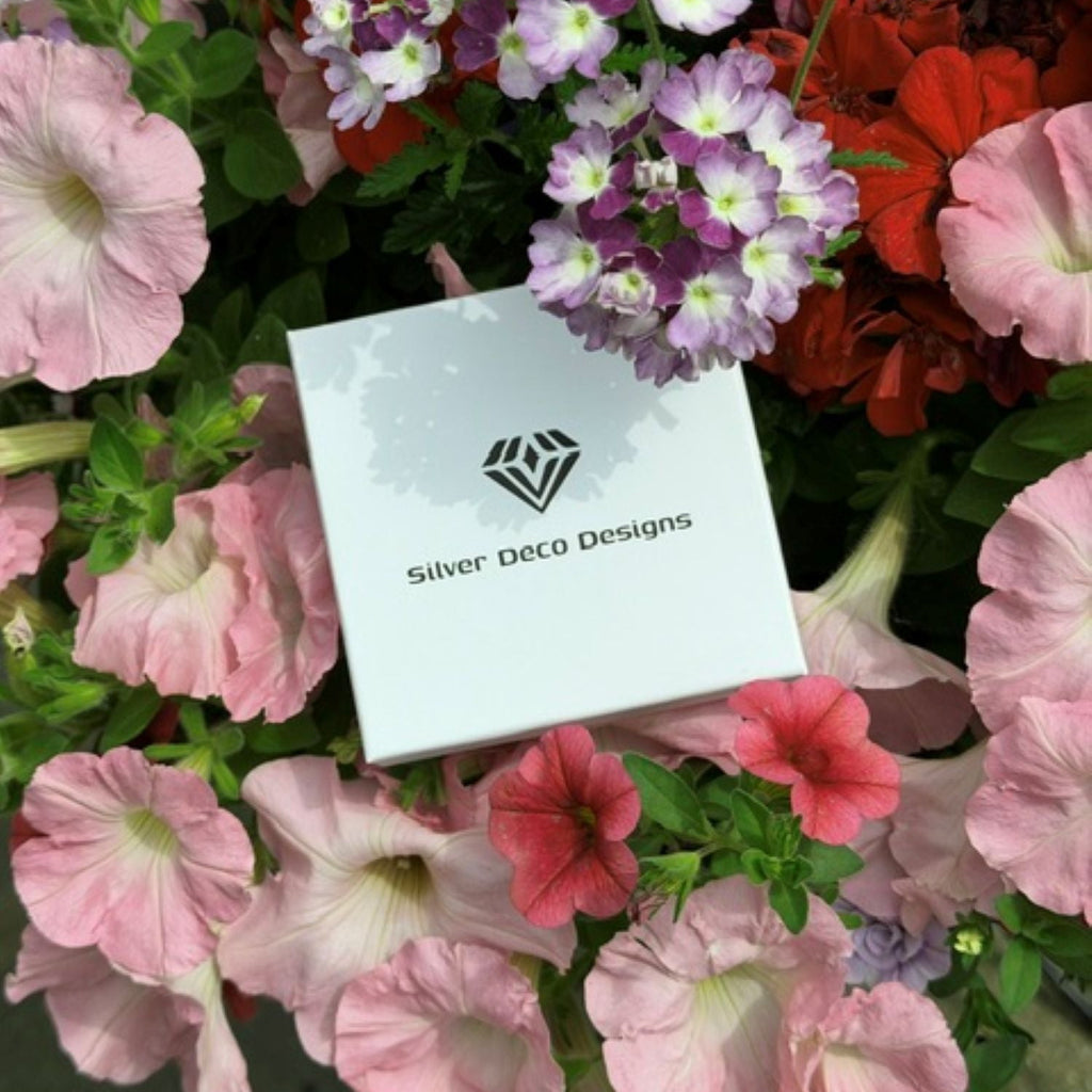 A white square jewellery box with the Silver Deco Designs logo and name, nestled among pink, red, and purple flowers with green foliage.