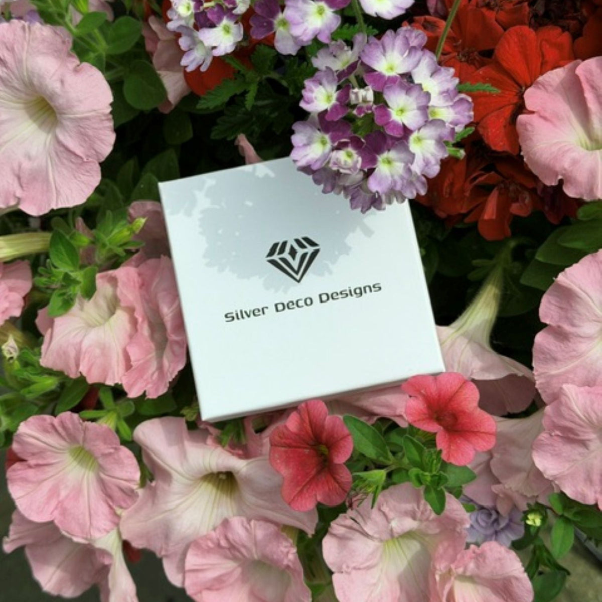 A white square jewellery box with the Silver Deco Designs logo and name, nestled among pink, red, and purple flowers with green foliage.