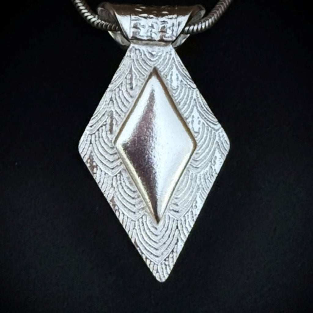 A textured silver pendant necklace featuring a central plain raised diamond shaped feature shown on a black background.