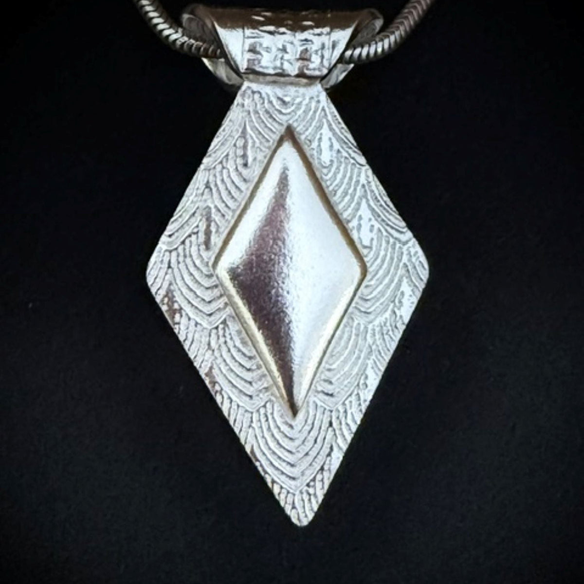 A textured silver pendant necklace featuring a central plain raised diamond shaped feature shown on a black background.