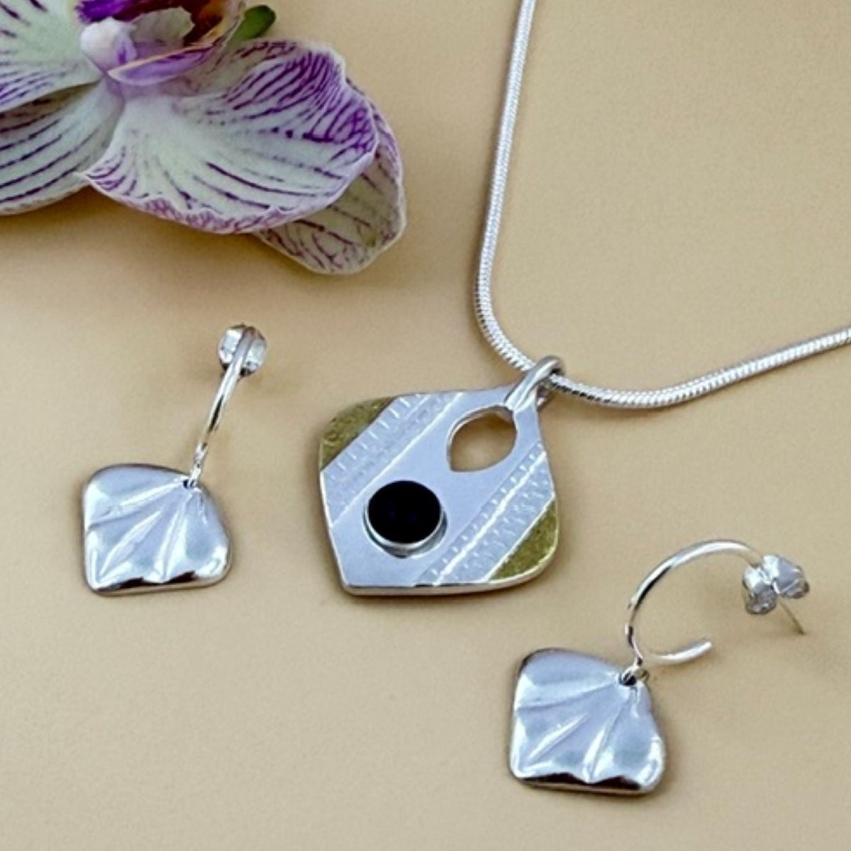 A jewellery set featuring a silver pendant necklace, matching drop earrings, all with textured elements, gold accents and black onyx gemstone. The set is displayed on a beige background next to a purple and white orchid flower.
