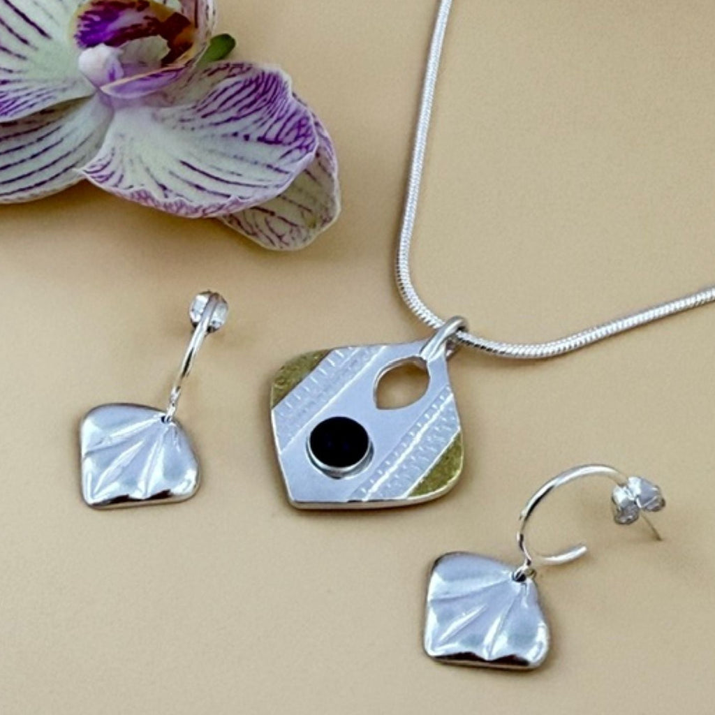 A jewellery set featuring a silver drop-shaped pendant necklace with added gold and a black onyx gemstone in the centre complemented by fan shaped drop earrings. The set is displayed on a beige background next to a purple and white orchid flower.
