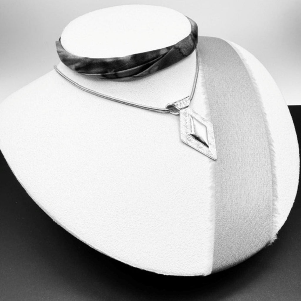 A silver geometric pendant necklace displayed on a white jewelry bust, with a dark patterned fabric choker also shown on the bust.