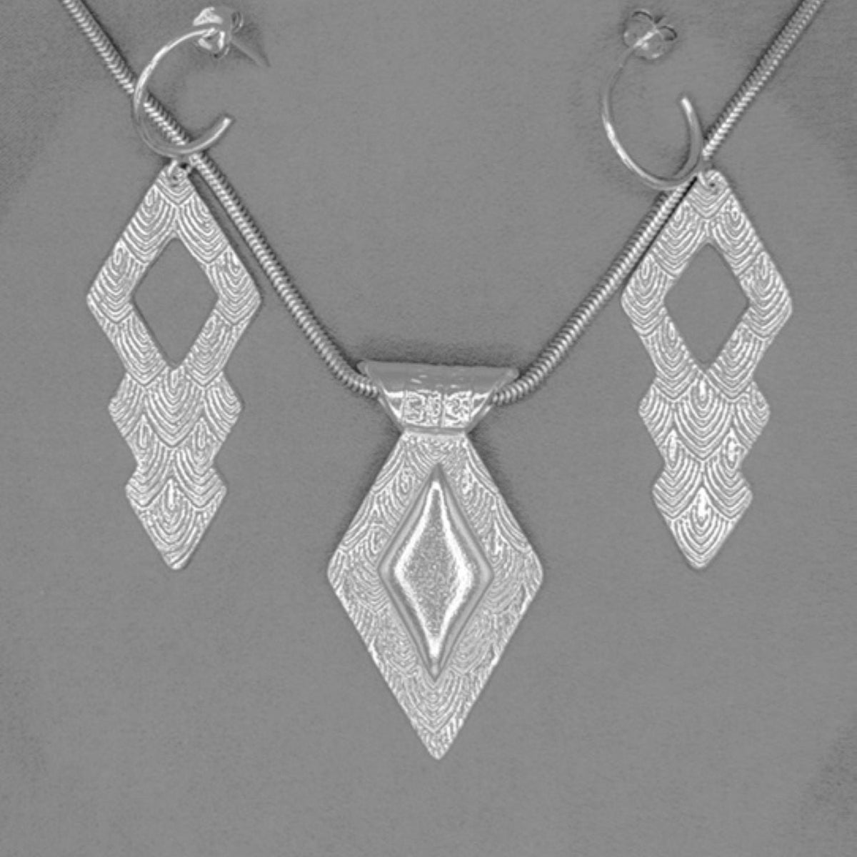 Silver jewelry set featuring a pendant necklace, matching drop earrings, and hoop earrings with textured geometric diamond designs, displayed on a neutral gray background.