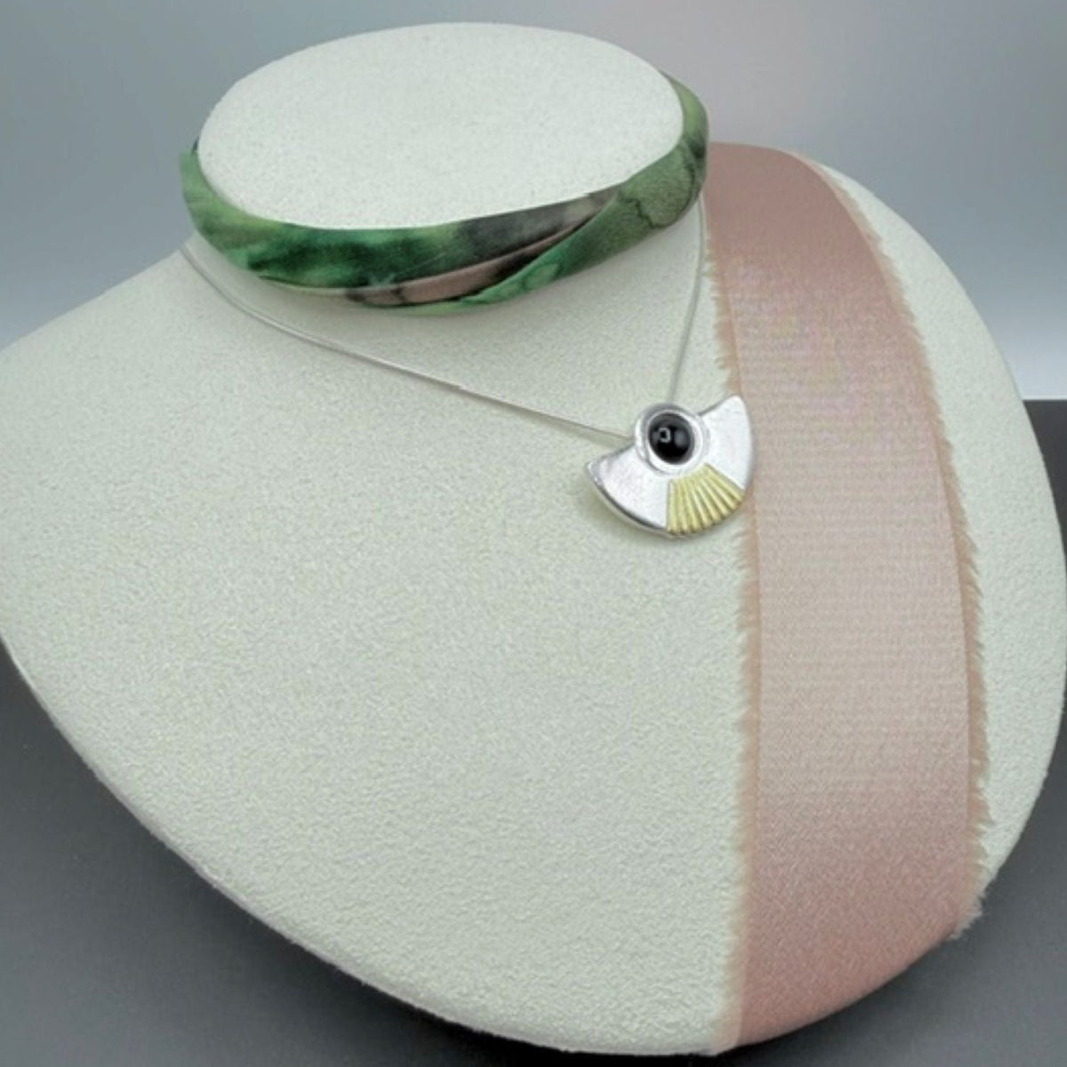 A silver pendant necklace with in a fan shaped design, featuring a black onyx gemstone and a gold sunburst pattern, displayed on a cream-colored jewellery bust