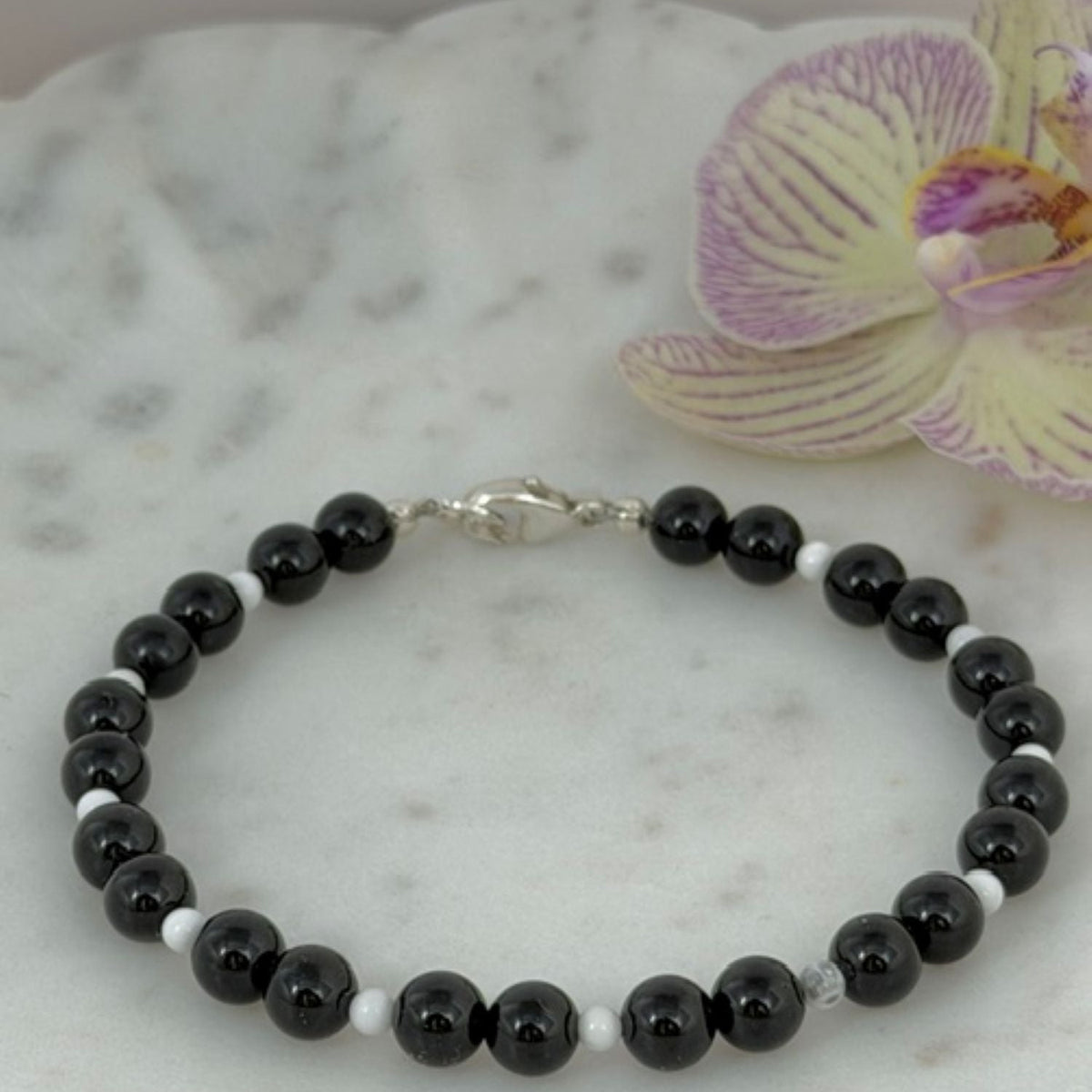 A beaded bracelet made of black onyx round beads separated by small howlite beads, with a silver clasp, displayed on a white marble surface next to a purple orchid flower.