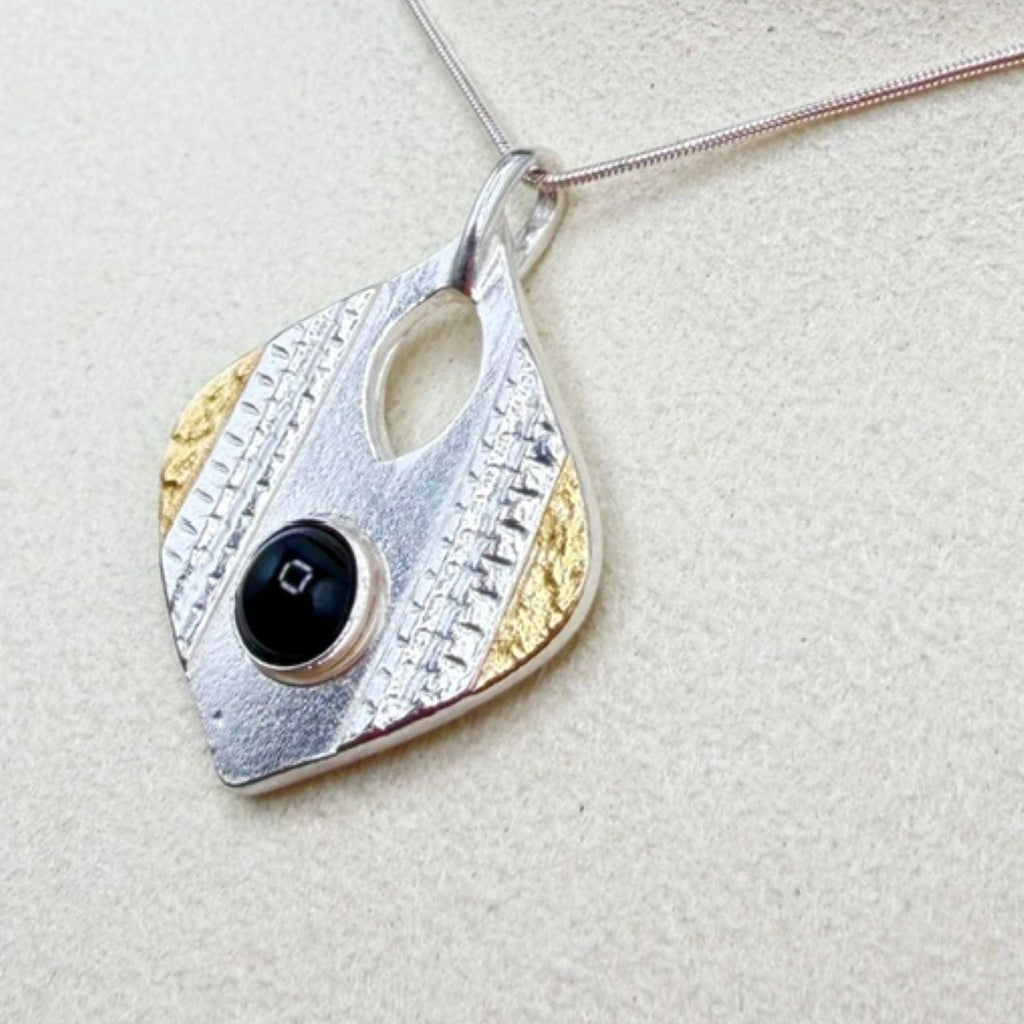 A silver and gold textured pendant necklace with a central black onyx stone, displayed on a textured beige surface.