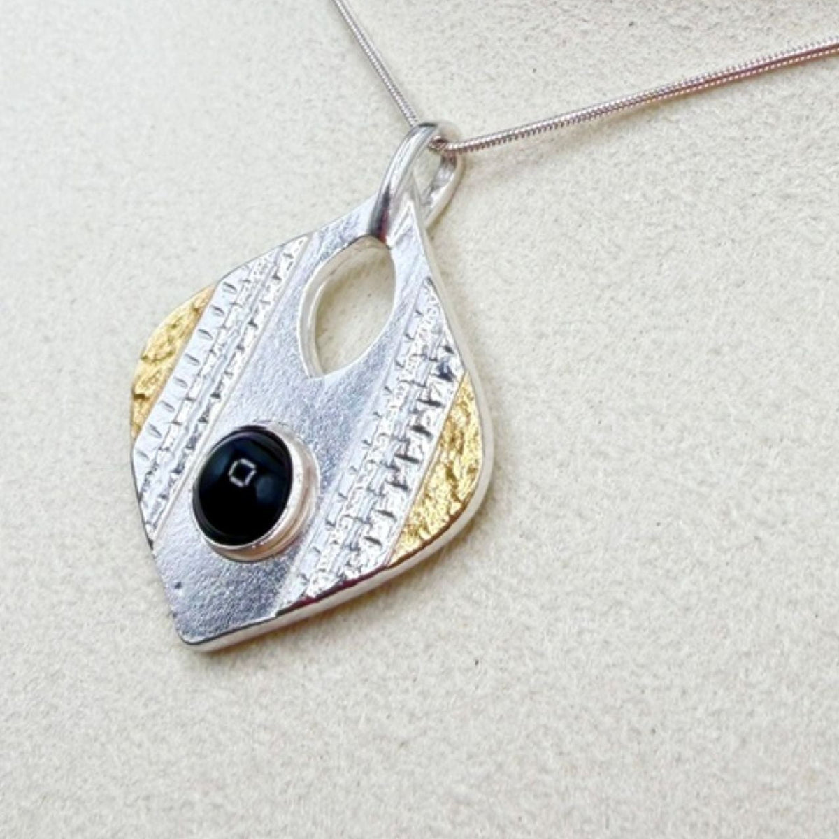 A silver and gold textured pendant necklace with a central black onyx stone, displayed on a textured beige surface.