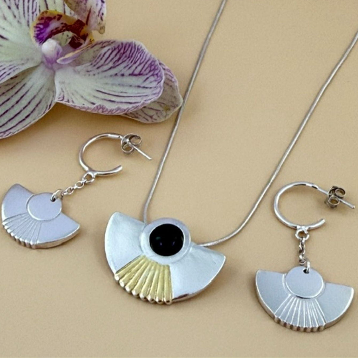 Silver fan-shaped jewellery set with a pendant necklace and a matching drop earring, featuring a black onyx gemstone and gold accent on a beige background with a decorative orchid.