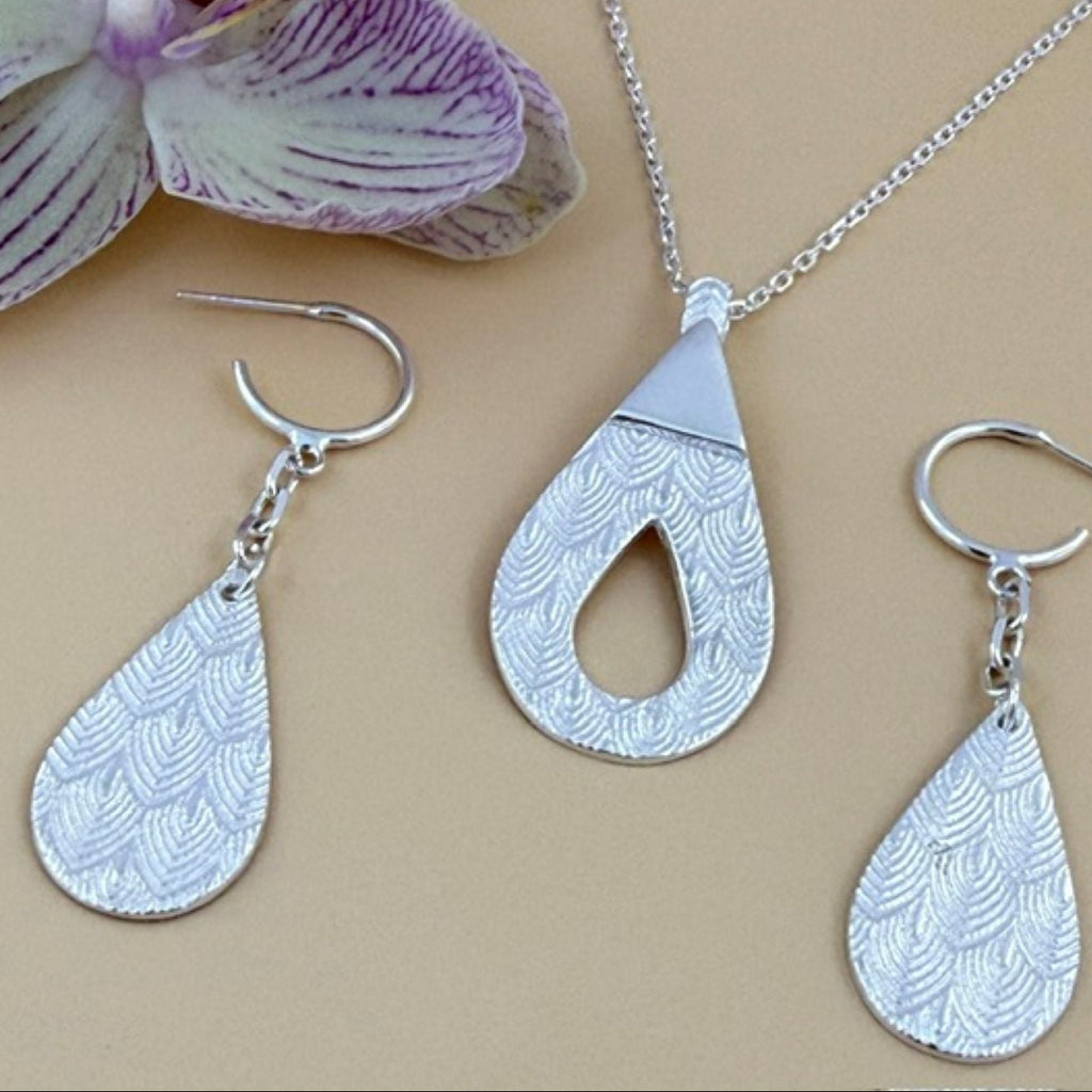 Silver jewelry set featuring a teardrop pendant necklace and two matching teardrop dangle earrings with a textured scallop pattern, displayed on a beige background next to a purple and white orchid flower.