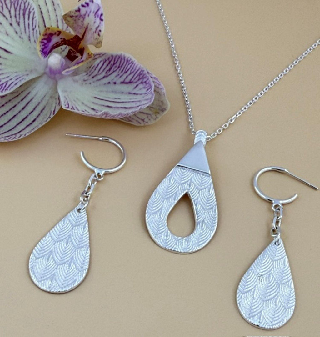 Silver jewelry set featuring a teardrop pendant necklace and two matching teardrop dangle earrings with a textured scallop pattern, displayed on a beige background next to a purple and white orchid flower.