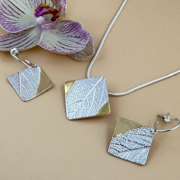 Silver jewellery set featuring a tilted square pendant necklace, matching drop earrings, all with textured silver leaf-patterned faces and gold accents, arranged on a beige background with a purple orchid flower.