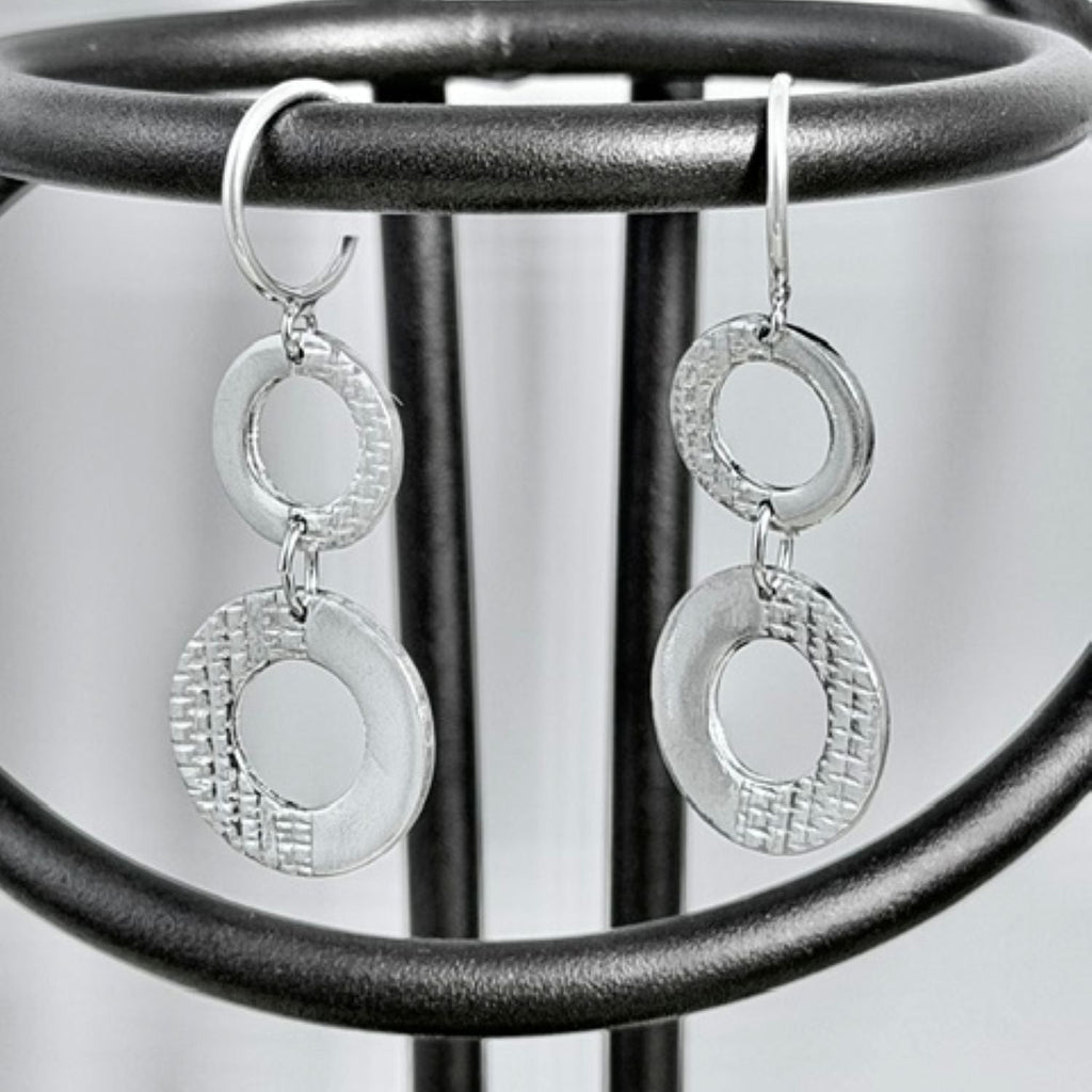 A pair of silver dangle earrings with textured circular disc elements and smooth oval links, hanging from a black metal display stand.