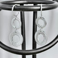 A pair of silver dangle earrings with textured circular disc elements and smooth oval links, hanging from a black metal display stand.