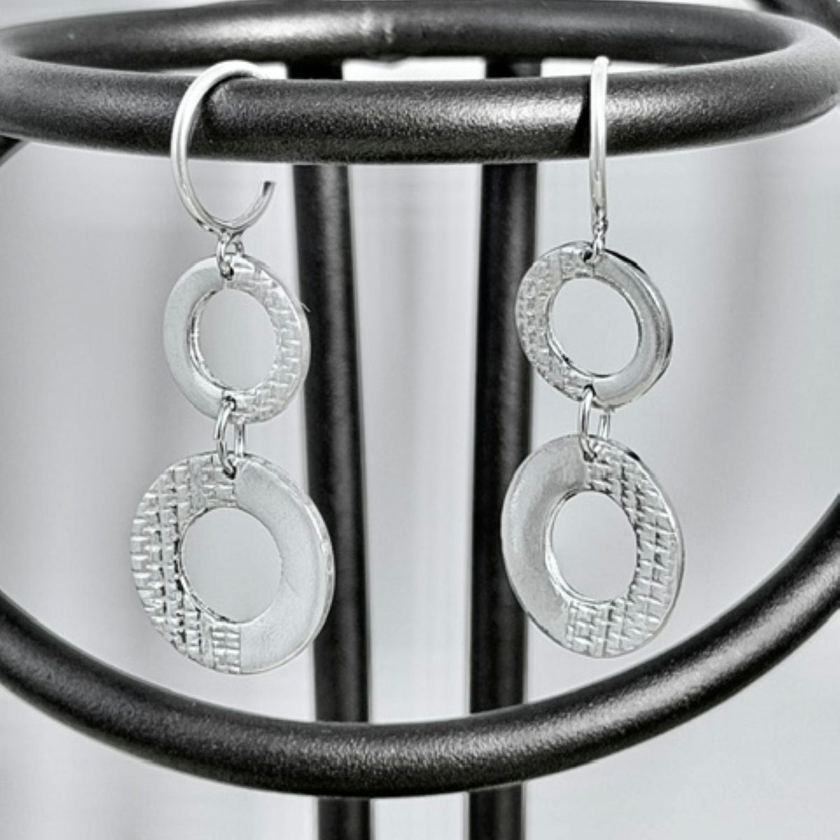 A pair of silver dangle earrings with textured circular disc elements and smooth oval links, hanging from a black metal display stand.