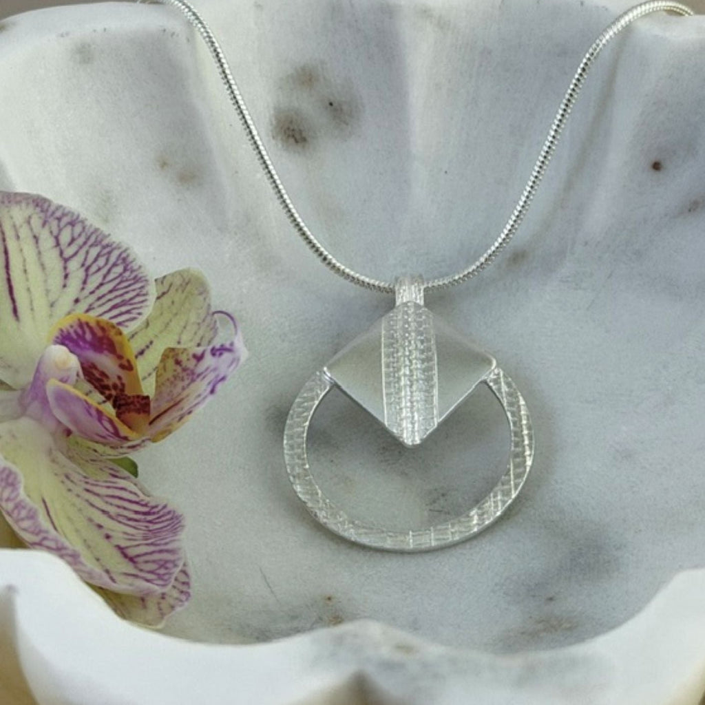 A silver circular pendant necklace showcasing a delicate texture with a tilted square feature, displayed on a white scalloped dish next to a purple and yellow orchid.