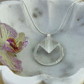 A silver circular pendant necklace showcasing a delicate texture with a tilted square feature, displayed on a white scalloped dish next to a purple and yellow orchid.