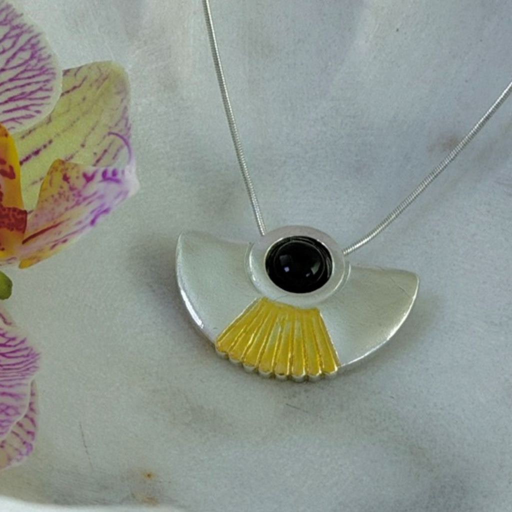 A silver pendant necklace with a fan-shaped design, featuring a textured yellow accent and a central onyx gemstone, displayed on a light surface next to a purple and yellow orchid flower.
