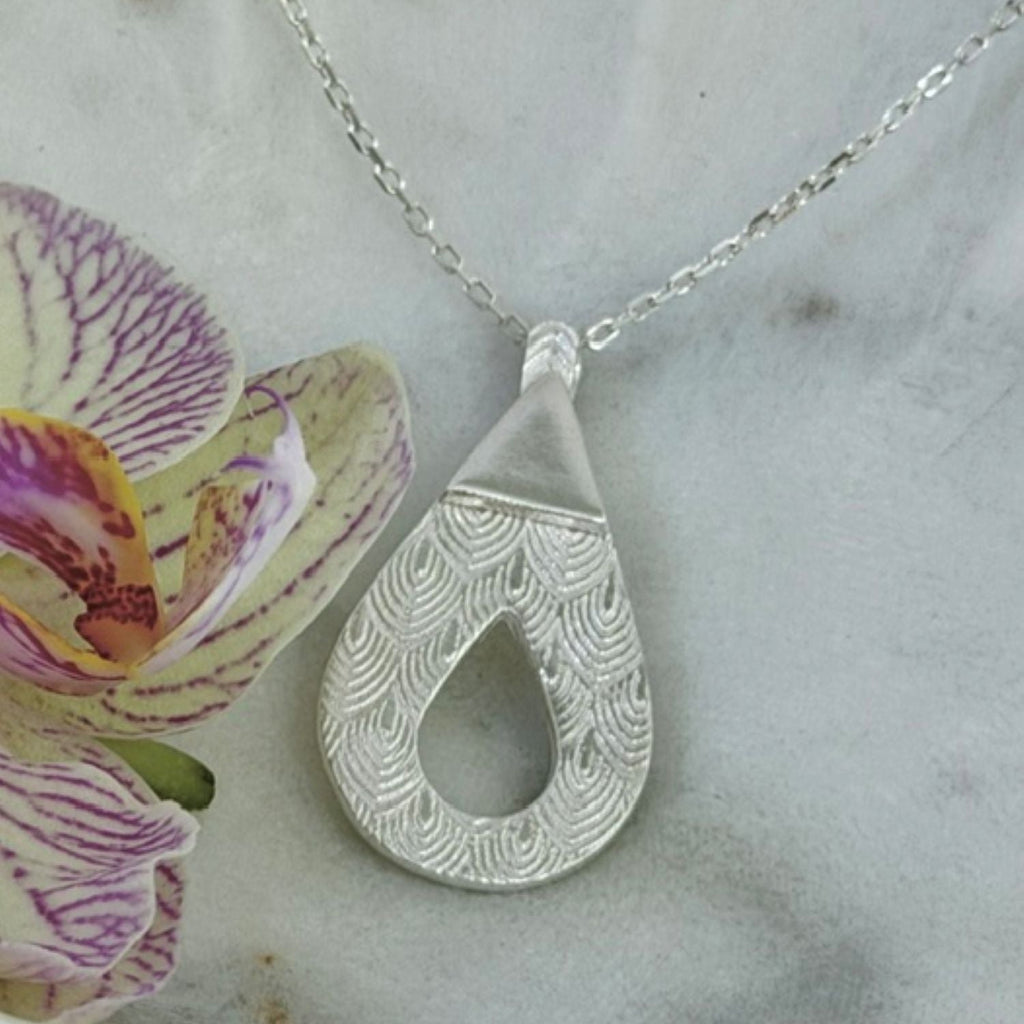 A silver teardrop pendant necklace with a textured scallop pattern, resting on a light grey marble surface next to a purple and yellow orchid flower.