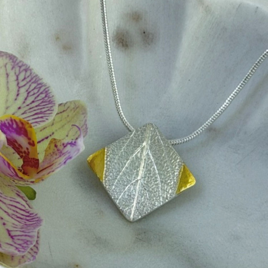A silver geometric pendant necklace with a textured leaf design and gold triangular side accents, resting on a white ceramic dish next to a purple and yellow orchid flower.