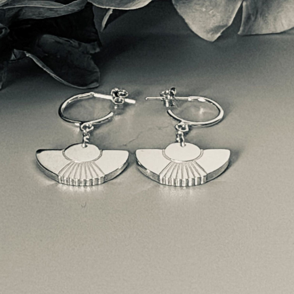 A pair of silver fan-shaped earrings, displayed on a neutral grey surface with dark dried leaves in the background.