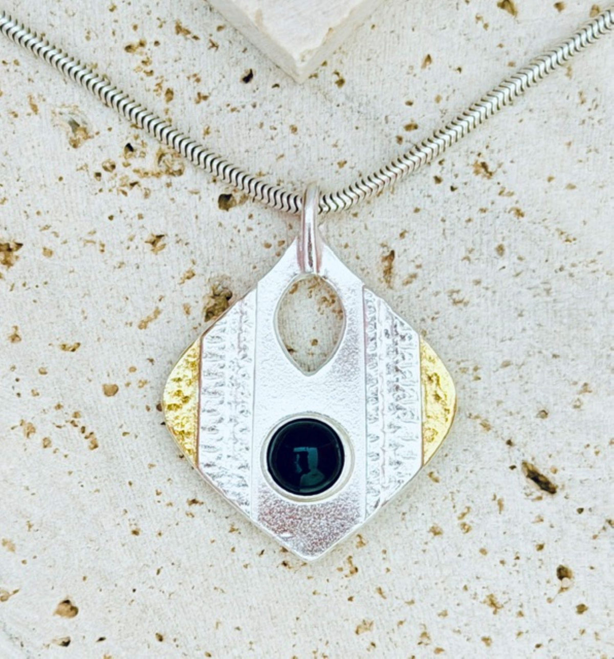 A textured silver pendant necklace with a diamond-shaped design, featuring a central oval cutout, a small round black gemstone, and gold-toned accents, displayed on a speckled stone surface.